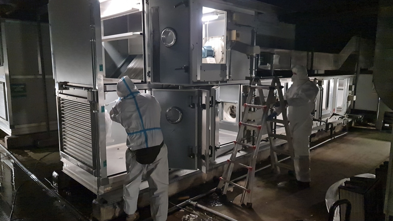 Intervento notturno su unità di trattamento aria — Rentacs, Service Key Group Due tecnici in tuta protettiva al lavoro di notte sulle sezioni aperte di un'unità di trattamento aria in copertura